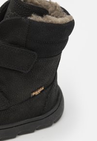 Bottine noire avec cuir texturé, doublure en laine douce et une sangle Velcro. Présente une semelle en caoutchouc robuste et un petit accent de marque.