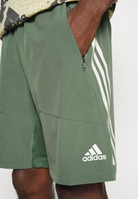 Vihreät urheilusortsit, joissa on valkoiset sivuraidat ja adidas-logo. Varustettu vetoketjullisilla taskuilla ja sileällä, kevyellä kankaalla.