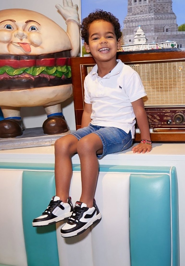 Zwart-witte sneakers met een rode accenten, denim shorts en een wit polo-shirt. Zittend naast een cartoon hamburger figuur.