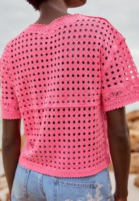Różowa, szydełkowana bluzka z krótkim rękawem w wzór diamentowej siatki z falbanką na dole, w połączeniu z jasnoniebieskimi, zniszczonymi szortami dżinsowymi.