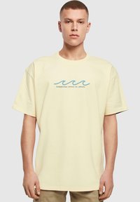 Gul bomulds T-shirt med løs pasform, der har et blåt bølgemotiv og teksten "lykke kommer i bølger" centreret under det.