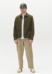 Jachetă verde olive cu închidere cu nasturi și două buzunare, purtată peste o cămașă albă și pantaloni bej; asortată cu adidași crem și o șapcă verde.