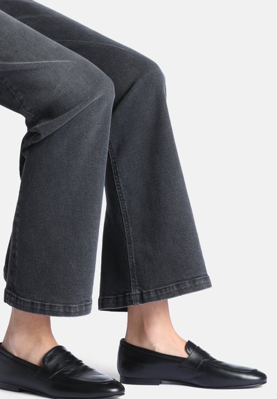 Jambes portant un jean noir à jambes larges et des mocassins en cuir noir sans lacets sur un fond clair uni.