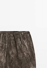 Pantalon marron en tissu texturé avec passants de ceinture et plis à la taille sur fond blanc uni.
