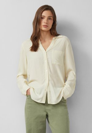 Frau mit langen braunen Haaren, die ein hellgelbes Hemd mit Knöpfen und olivgrüne Hosen trägt, steht mit den Händen in den Taschen vor einem grauen Hintergrund.