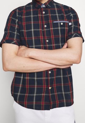 Mann mit verschränkten Armen, der ein kurzärmeliges marineblau kariertes Hemd mit roten, gelben und weißen Linien und einer Brusttasche mit Knopfdetail trägt.