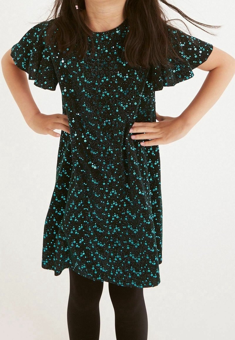 Robe noire avec un motif de sequins teal, manches courtes évasées et taille cintrée, accompagnée de leggings noirs. La texture est lisse et brillante.