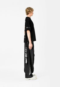 Camiseta negra de gran tamaño con estampado gráfico en la espalda, combinada con pantalones oscuros holgados que tienen un zipper lateral y zapatos blancos de suela gruesa.