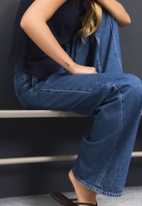 Vidbenede denimjeans i en mediumblå vask med kontrastsyning og en afslappet pasform, kombineret med en navyblå top og brune sandaler.