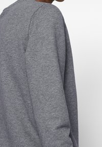 Sudadera gris con cuello redondo y mangas largas, hecha de un tejido suave y texturizado, con un corte relajado. Diseño sencillo sin patrones visibles.