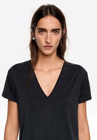 Czarny t-shirt z krótkim rękawem i dekoltem w serek, o gładkiej teksturze, ozdobiony małymi metalicznymi ćwiekami ułożonymi w kropkowany wzór na całej powierzchni.