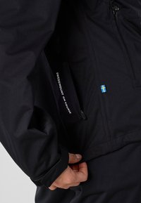 Main ajustant le poignet d’une veste noire avec une poche zippée et un petit écusson du drapeau suédois sur la poitrine.