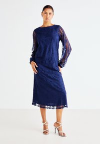 Vestido de renda azul-marinho com mangas longas transparentes, padrão floral e corte a meio comprimento. Acompanhado de saltos altos prateados com tiras.
