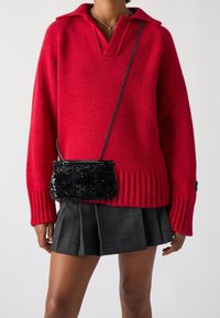 Persona che indossa un luminoso maglione rosso a maglia, una gonna plissettata grigia a quadretti e una piccola borsa a tracolla nera con paillettes, dotata di tracolla a catena.