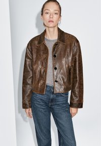 Bruine leren cropped jack met een klassieke kraag, een sluiting met knoppen aan de voorkant en zijzakken. Draag het over een grijs t-shirt en blauwe jeans.