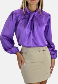 Blusa di raso viola con scollo annodato e maniche a sbuffo, abbinata a una gonna a matita beige con due tasche decorative e dettagli con bottoni.