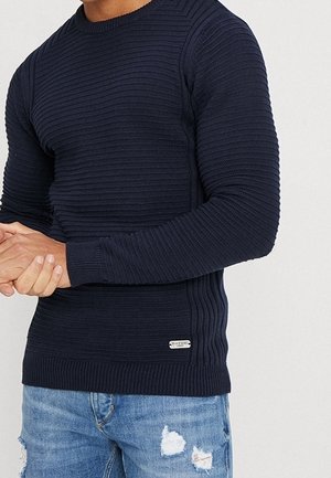 Un homme portant un pull en maille côtelée bleu marine et un jean bleu déchiré, les mains jointes devant lui, sur un fond clair uni.