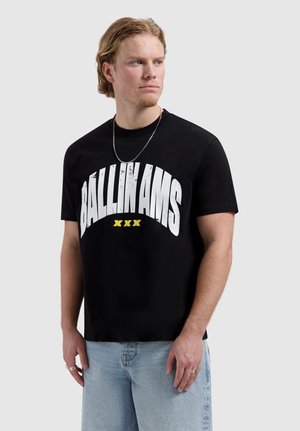 Man met blond haar, gekleed in een zwart T-shirt met "BALLIN AMS XXX" in wit en geel, lichtblauwe spijkerbroek en een zilveren ketting, kijkt naar links.