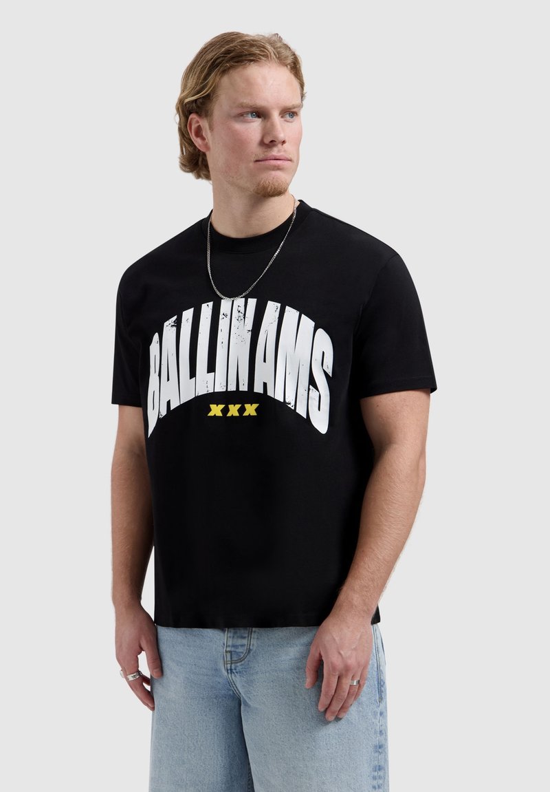 Man met blond haar, gekleed in een zwart T-shirt met "BALLIN AMS XXX" in wit en geel, lichtblauwe spijkerbroek en een zilveren ketting, kijkt naar links.