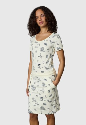 Kvinde iført en cremefarvet kjole med korte ærmer og sorte sommerlige illustrationer, stående med den ene hånd i lommen.