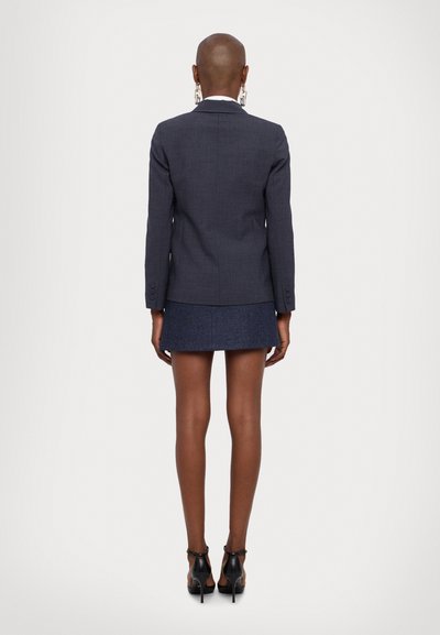 Blazer navy su misura con finitura testurizzata, forma aderente e chiusura con bottone singolo; abbinato a una mini gonna e scarpe col tacco nere.