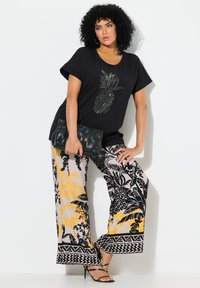 Černé tričko s designem ananasu, kombinované s volnými kalhotami s vzorem v žluté, černé a béžové barvě. Drží černou kabelku.