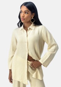 Camicia button-up color crema chiaro con tessuto strutturato e sgualcito. Maniche lunghe, vestibilità rilassata e davanti aperto che mostra i pantaloni coordinati.