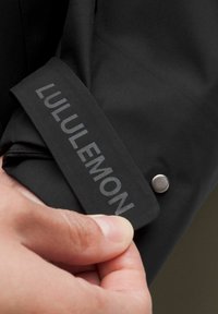 Sort jakkeærme med sølv snaps og "LULULEMON" trykt med refleksiv tekst på et glat, mat stof.