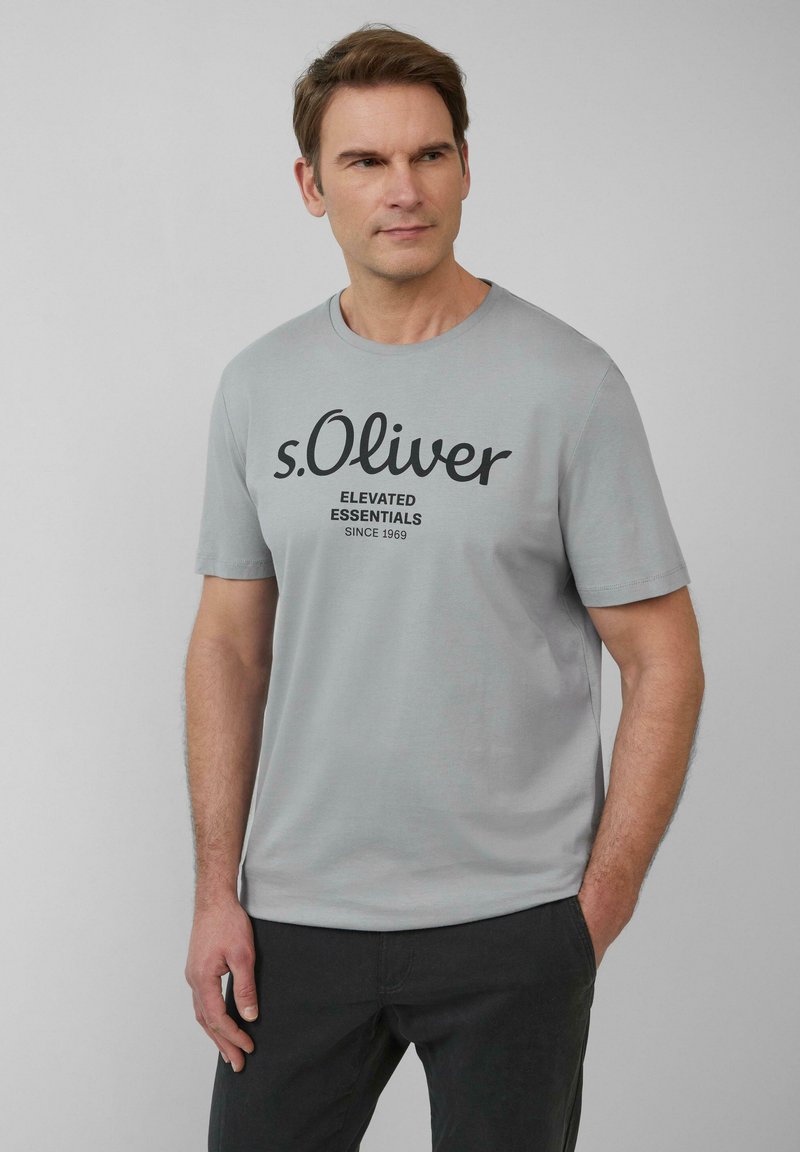 Jasnoszara koszulka z krótkim rękawem z czarnym nadrukiem tekstu "s.Oliver" oraz "ELEVATED ESSENTIALS SINCE 1969." Wykonana z miękkiego materiału z okrągłym dekoltem.