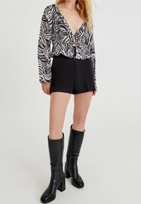 Musta-valkoinen seepra-kuosinen paita, jossa röyhelöitä ja syvä v-pääntie, yhdistetty korkeavyötäröisiin mustiin shortseihin ja polven korkeisiin mustiin korkosaappaisiin.