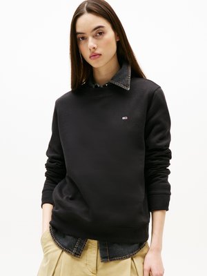 Mustapaita, jossa on ribbireunat hihassa ja helmassa, ja pieni logo. Paita on sinisen farkkutakin päällä, jossa on näkyvissä oleva kaulus. Khaki-housut.