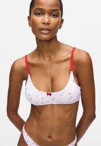 Moteris vilki baltą liemenėlę ir kelnaites su mažais raudonais širdelėmis ir raudonomis juostelėmis prieš paprastą baltą foną.