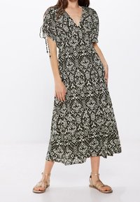 Maxikleid in dunkelgrün mit weißen geometrischen Mustern, V-Ausschnitt, kurzen Ärmeln, Kordelzugdetails und einem fließenden Saum. Flache goldene Sandalen getragen.