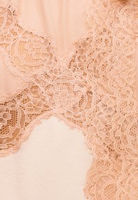 Λεπτό δαντελένιο ύφασμα ροδάκινου με λουλουδένια σχέδια, στρωμένο πάνω σε λείο ύφασμα ροδάκινου, παρουσιάζοντας περίτεχνη κεντητική και κυματιστά άκρα.