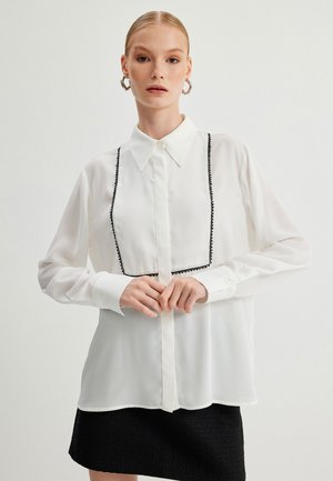 Blusa blanca de manga larga con cuello, que presenta un ribete decorativo negro a lo largo del frente, combinada con una falda negra texturizada.