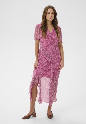 Vrouw draagt een roze bloemen wikkeljurk met gerimpelde zoom en korte mouwen, staand met bruine blokhak schoenen op een witte achtergrond.