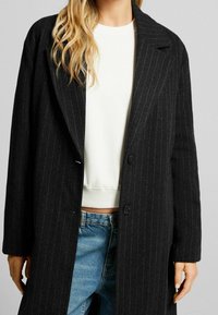 Cappotto a righe nere realizzato in tessuto testurizzato, con colletto a punta, due bottoni e maniche morbide, indossato sopra una maglia bianca.