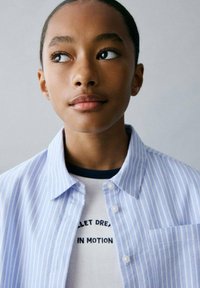 Cămașă cu dungi albastre deschise, cu guler, suprapusă peste un tricou alb cu text negru, cu mâneci scurte și gât rotund.