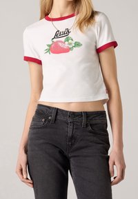 Camiseta blanca recortada con cuello rojo y gráfico grande de una fresa, con el texto "Levi's" arriba. Combinada con jeans oscuros.