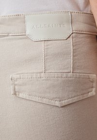 Vaqueros de mezclilla beige que presentan un parche de cuero con el logotipo "ALLSAINTS" en relieve, costuras en el bolsillo trasero y una textura suave.