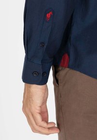 Chemise à manches longues bleu marine avec des manchettes à boutons, arborant un logo de cheval rouge sur la manche et un accent rouge contrastant à l'ourlet.