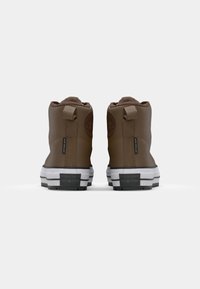 Brown high-top sneakers with a textured finish, contrasting white midsole, and rear pull tab featuring a logo patch. Black rubber outsole.