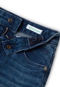 Vaqueros de denim en un lavado azul oscuro con un corte slim. Presenta cremallera de metal, botones y una etiqueta verde que dice "BRITNEY-SLIM." Detalle notable de costura.