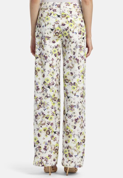 Femme portant un pantalon taille haute à jambes larges, avec une base blanche et un imprimé floral jaune et violet, debout avec les bras détendus sur les côtés.