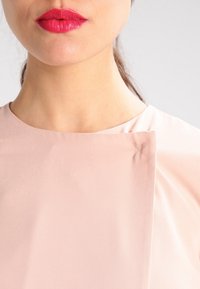 Indumento di colore rosa chiaro con una texture liscia e un colletto rotondo. Un dettaglio notevole include una piccola cucitura sul tessuto superiore.