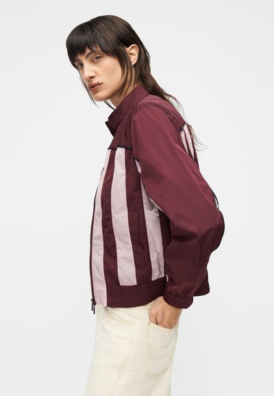 Kvinde med mørkt hår iført en vinrød og lyserød stribet jakke og cremefarvede bukser, står afslappet mod en ensfarvet baggrund.