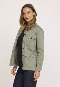 Veste vert clair en coton avec fermeture à boutons, deux poches poitrine et poignets à boutons. Portée par-dessus un haut noir et associée à un jean foncé.