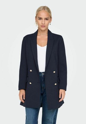 Marineblå dobbeltknappet blazer lavet af glat stof med guldknapper, der har spidse klapper og sidelommer, båret over en hvid top.
