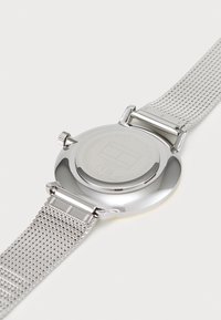 Reloj de malla plateada con una caja redonda reflectante y una correa de metal texturizada. Presenta una corona y un logo grabado en la parte posterior.