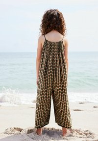 Mørkegrøn blomsterjumpsuit med tynde stropper, sammensamlet bryststyke og brede ben, båret af et barn, der står på stranden med ansigtet mod havet.